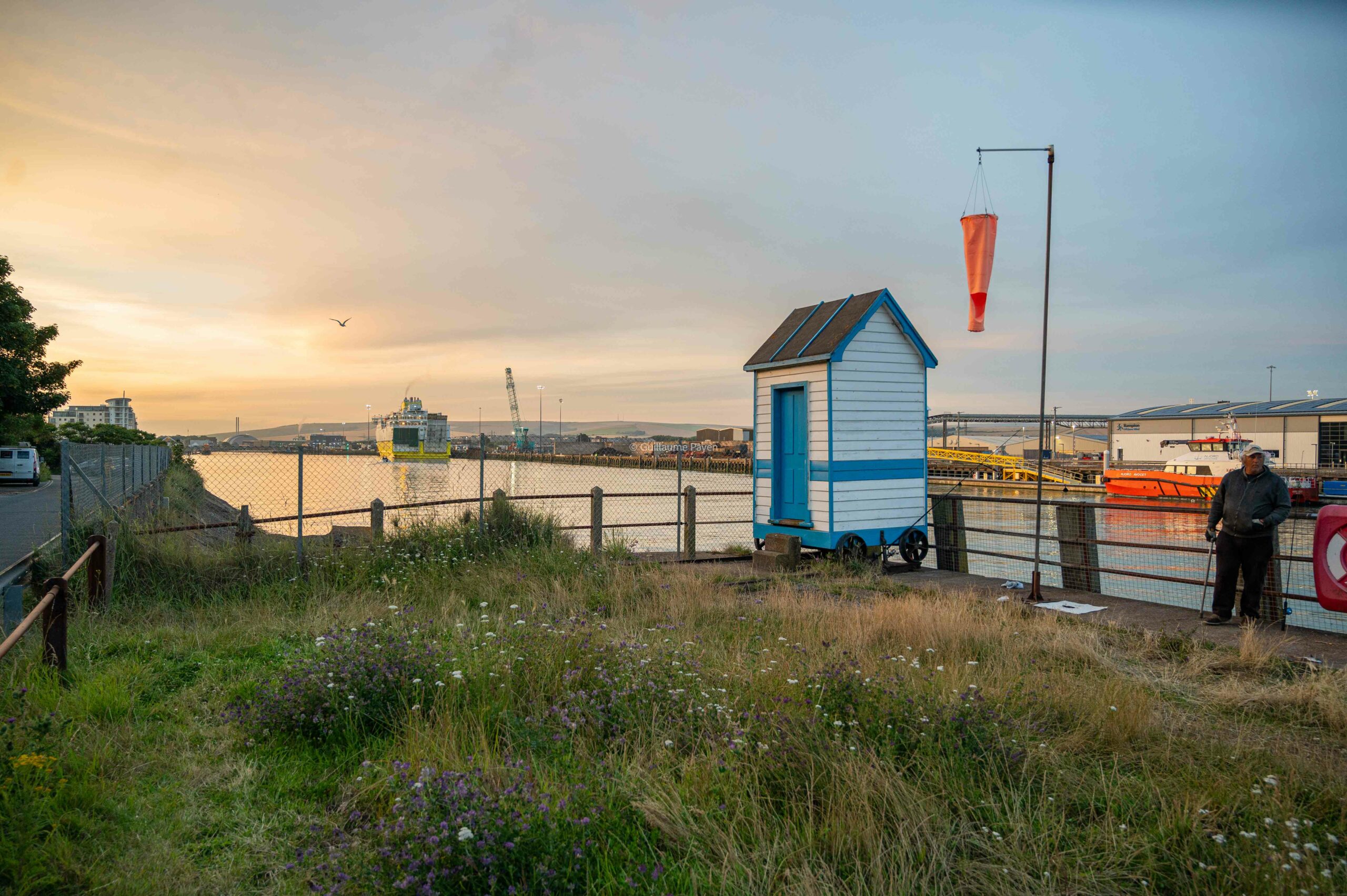 On voit en premier plan une cabane de pêcheur avec un pecheur au près, un rivière et au loin le ferry jaune qui part pour la Normandie 